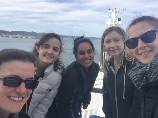 Five women marine scientists stop for a photo on the bow of RV Investigator while the ship is fairly close to the coastline (visible in the background) before heading out on the Tasman Sea to collect pore water for chromium and neodymium analysis.