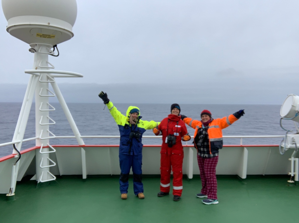 Three female scientists stand on an outside deck area in heavy water proof gear taking a break from observations to pose for a picture.